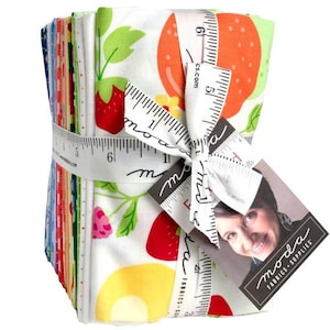 May include: A stack of colorful fabric squares tied with a white ribbon printed with ruler markings and the word "moda". The fabric features a fruit pattern with strawberries, lemons, and oranges. A tag with a woman's image is attached.