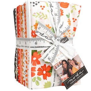 May include: A stack of folded fabric squares tied with a white ribbon printed with a black measuring tape design and the text Moda Fabrics. The fabric features floral and geometric patterns in orange, white, green, and black. A tag shows a photo of two people.