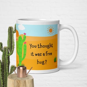 May include: White ceramic mug with a desert scene featuring a green cactus, orange sand, blue sky, and a sun. The mug has the text "You thought it was a free hug?" in black. A copper watering can sits in front of the mug.
