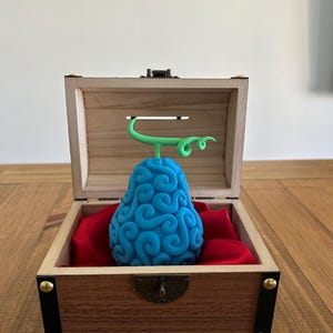 Peut inclure: Un fruit bleu en forme de cerveau avec une tige verte repose sur un tissu de satin rouge à l'intérieur d'un coffre au trésor en bois. Le coffre est ouvert, révélant le fruit, et est décoré d'éléments métalliques.