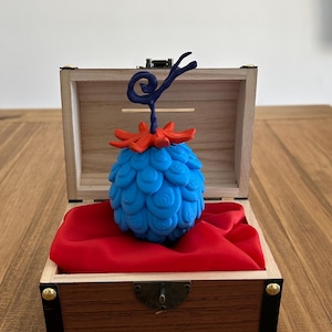 Peut inclure: Une sculpture bleue en forme de fruit avec une base rouge et une tige bleu foncé, présentée dans un coffre au trésor en bois ouvert, doublé de tissu rouge. Le fruit a un motif tourbillonnant texturé.