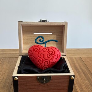 Peut inclure: Un objet rouge en forme de cœur avec un motif tourbillonnant et une tige turquoise repose sur un tissu noir à l'intérieur d'une boîte en bois ouverte. La boîte a un couvercle à charnière et une base marron foncé avec des accents dorés.