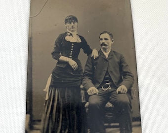 May include: A black and white tintype photograph of a couple posing for a portrait. The woman is wearing a long dress with a lace collar and the man is wearing a suit and tie. They are both looking at the camera.