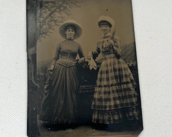 May include: A black and white tintype photograph of two women wearing hats and dresses. The woman on the left is wearing a dark dress and a wide-brimmed hat. The woman on the right is wearing a plaid dress and a smaller hat. They are standing in front of a tree and a fence.
