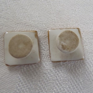 May include: Two square ceramic knobs with a gold trim and a white center. The knobs have a round, white base.