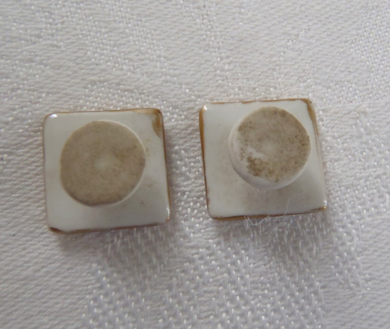 May include: Two square ceramic knobs with a gold trim and a white center. The knobs have a round, white base.
