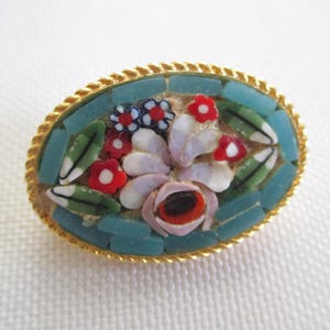 May include: Oval mosaic brooch with a gold-tone rope edge. The mosaic features a floral design with red, white, and blue flowers, green leaves, and a light blue background. A vintage-style accessory.
