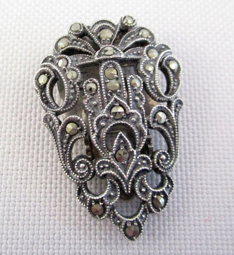 Puede incluir: Un broche plateado con un dise&ntilde;o floral detallado, engastado con piedras de marcasita.
