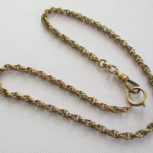 May include: A gold-toned watch chain with a detailed, woven design. The chain features a lobster clasp and a small ring at the end. The chain is displayed on a white surface, showcasing its intricate links and vintage aesthetic.