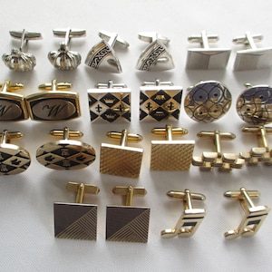 May include: A collection of vintage cufflinks in various designs. Styles include gold-toned and silver-toned metal with geometric, patterned, and initialed faces. Some feature crown, shield, and abstract shapes. A variety of shapes, including square, oval, and round.