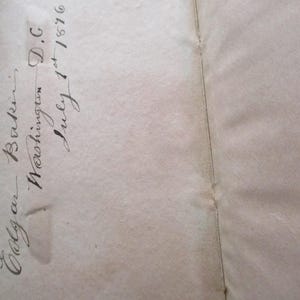 May include: Open book pages with handwritten script. The script reads "Edgar Baker, Washington D.C. July 1st 1876". The paper is aged with a light brown colour and a vertical seam.