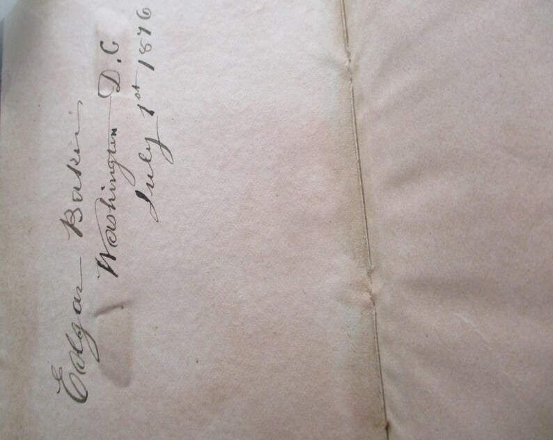 May include: Open book pages with handwritten script. The script reads "Edgar Baker, Washington D.C. July 1st 1876". The paper is aged with a light brown colour and a vertical seam.