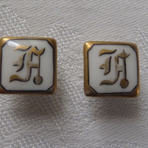 May include: A pair of gold-toned cufflinks with a white porcelain center featuring a gold-colored letter 'F' in a stylized font.