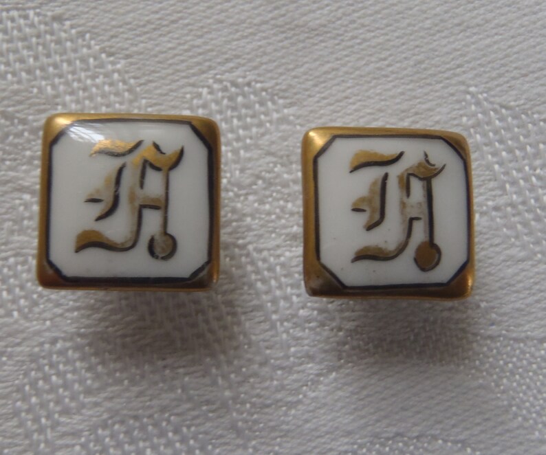 May include: A pair of gold-toned cufflinks with a white porcelain center featuring a gold-colored letter 'F' in a stylized font.