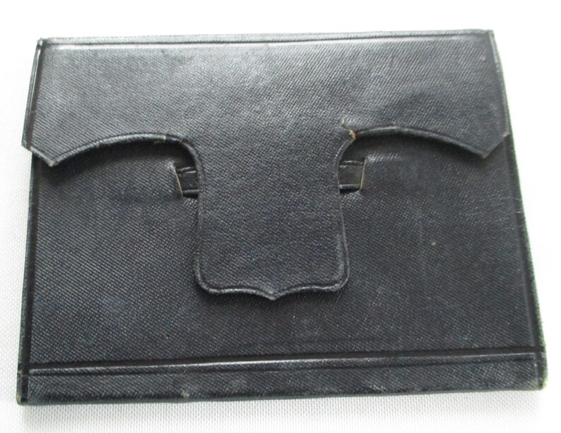 May include: A black rectangular case with a textured surface. It features a flap closure with a curved design and a small tab. The case is likely made from a durable material.