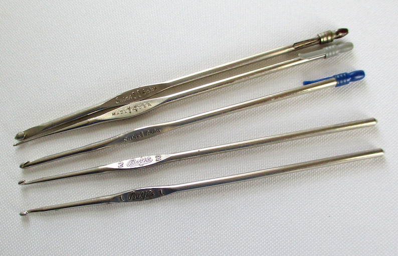 May include: Five silver crochet hooks of different sizes. The hooks are labeled with their sizes: "Clover 15/0", "Made in USA", "Size 14/0", "Clover 2", and "Clover 1".