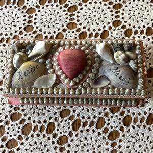 May include: Rectangular box decorated with seashells, pearls, and a heart-shaped element. The box features the word "Revere" on one shell and "Beach" on another. The box is trimmed with pink and sits on a lace surface.
