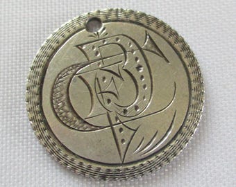 May include: A silver-colored, round pendant with an engraved design. The pendant features a stylized letter "B" with decorative elements and a textured border. A small hole is at the top for hanging. The pendant is likely made of metal.