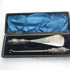 May include: A silver-toned shoehorn and a button hook set with ornate, embossed handles featuring intricate scrollwork. The two items are presented inside a rectangular black case lined with dark blue velvet and a blue satin interior lid.