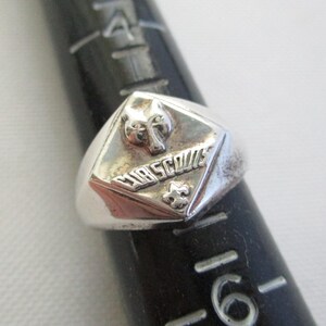 May include: A silver-toned ring with a diamond-shaped face featuring a wolf's head, the word "CUBSCOUTS", and a fleur-de-lis emblem. The ring is set on a black ring sizer, showing a size 16.