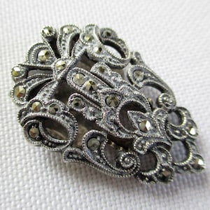 Puede incluir: Una pinza para el pelo plateada con un dise&ntilde;o detallado y ornamentado. La pinza est&aacute; cubierta de peque&ntilde;as piedras de marcasita brillantes.