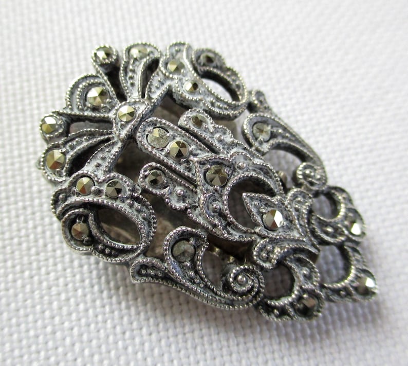 Puede incluir: Una pinza para el pelo plateada con un dise&ntilde;o detallado y ornamentado. La pinza est&aacute; cubierta de peque&ntilde;as piedras de marcasita brillantes.