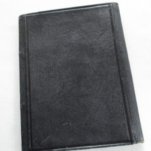 May include: A small, rectangular, black book with a textured cover. The book has a simple, classic design with a dark, solid colour. The edges of the book are slightly worn, suggesting age or use.
