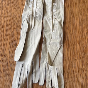 May include: A pair of long, light beige gloves featuring four small white buttons along the wrist area of each glove. The gloves have a smooth texture and extend past the wrist, with visible stitching along the fingers and a side opening detail.