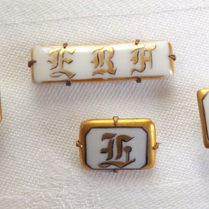May include: Four gold-rimmed porcelain buttons with the letter "F" in a stylized font. The buttons are white with gold lettering.