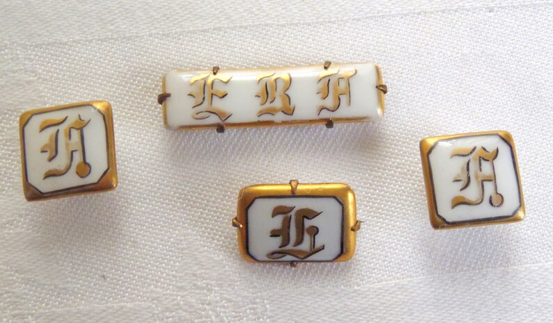 May include: Four gold-rimmed porcelain buttons with the letter "F" in a stylized font. The buttons are white with gold lettering.