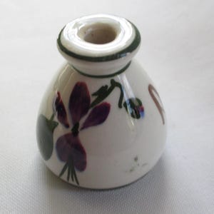 May include: A small, white ceramic candle holder with a rounded base and a circular opening at the top. The candle holder is decorated with a purple flower and green leaves. A green band encircles the top rim.