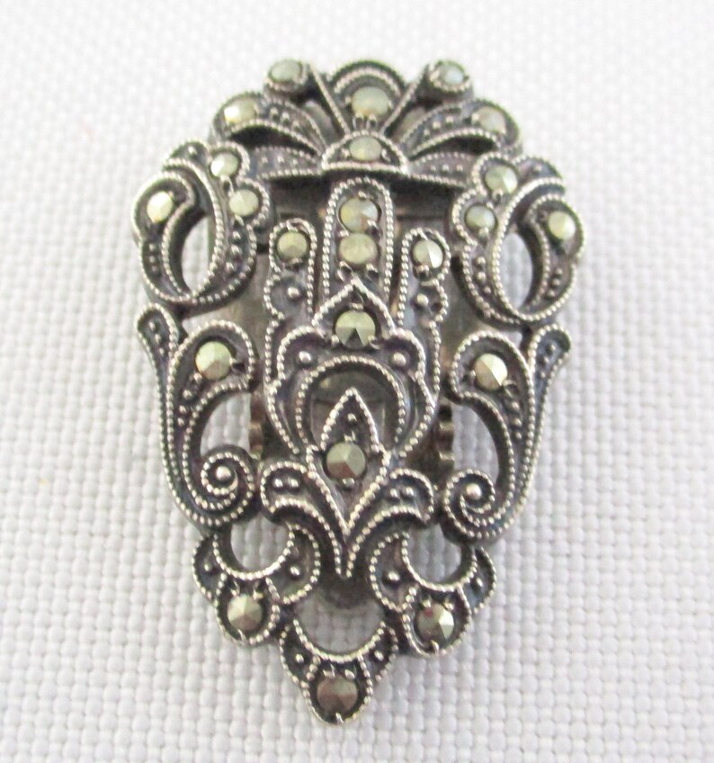 Puede incluir: Un broche de tono plateado con un dise&ntilde;o floral detallado, engastado con piedras de marcasita.