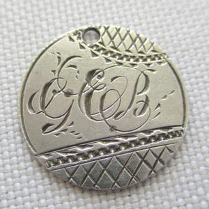 May include: A silver-colored, round pendant with a hole at the top. The pendant features engraved script letters "G C B" in the center, surrounded by decorative patterns. The edges have a diamond-shaped design.