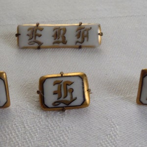 May include: Four gold-rimmed porcelain buttons with the letter "F" in a stylized font. The buttons are white with gold lettering and have a gold rim.