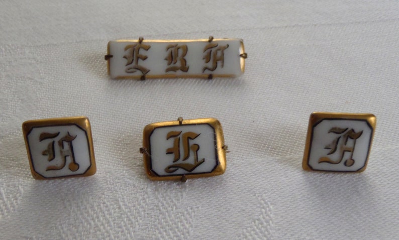 May include: Four gold-rimmed porcelain buttons with the letter "F" in a stylized font. The buttons are white with gold lettering and have a gold rim.