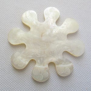 May include: A cream-colored, flower-shaped decorative piece with etched floral and circular designs. The piece has a pearlescent sheen and a unique, multi-lobed shape. The etched details include flowers and a central circular pattern.