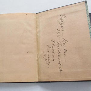 May include: An open antique book with aged, tan pages. The right-hand page features handwritten cursive text, including the name "Baker" and an address. The book has a black cover with a decorative green and white pattern along the edge.