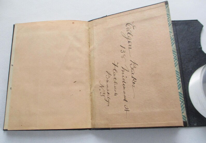 May include: An open antique book with aged, tan pages. The right-hand page features handwritten cursive text, including the name "Baker" and an address. The book has a black cover with a decorative green and white pattern along the edge.