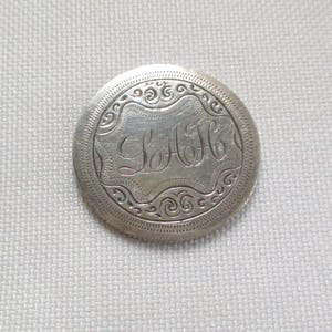 May include: A round, silver-toned button with an engraved design. The button features the initials "LAH" in an ornate script within a decorative border. The outer edge has a detailed, repeating pattern.