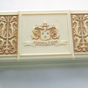 May include: Ivory-colored rectangular box with a decorative design. The box features a central panel with a crest and the name "HAMILTON" on a banner. Flanking panels have ornate, symmetrical patterns. The box has a slightly raised, textured surface.