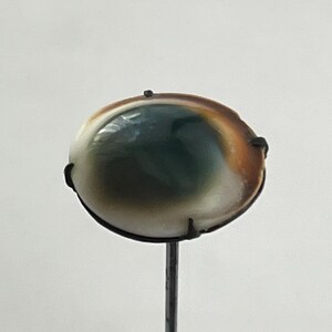 May include: An oval-shaped cabochon stone featuring a deep green center that transitions into a creamy white and warm brown outer edge. The stone is secured in a dark metal prong setting mounted on a thin, vertical metal pin or stem.