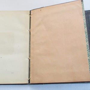 May include: An open, vintage book with cream and tan pages. The pages are bound with thread. The cover is dark grey with a decorative edge. The book is displayed open, revealing the interior pages.