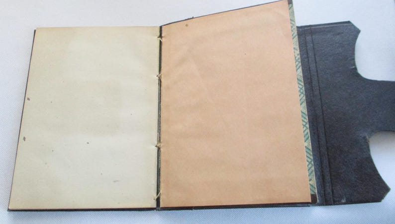 May include: An open, vintage book with cream and tan pages. The pages are bound with thread. The cover is dark grey with a decorative edge. The book is displayed open, revealing the interior pages.