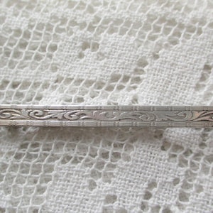 May include: A silver-toned tie bar with an ornate, symmetrical floral design. The rectangular bar features a detailed pattern of scrolling leaves and vines, adding a touch of vintage elegance. The bar is displayed on a white lace background.