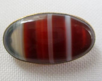 May include: A gold-toned oval brooch with a red, white, and gray banded agate stone.