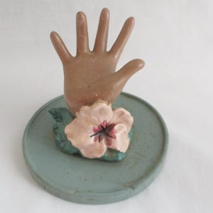 May include: A ceramic hand-shaped ring holder in a light brown color, with a pink flower and green leaves on a round, light blue base. The ring holder is a decorative item.