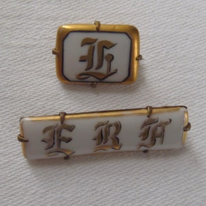 May include: Two gold-rimmed porcelain brooches with black and gold lettering. The first brooch is square and features the letter "F" in a stylized font. The second brooch is rectangular and features the letters "F R M" in a stylized font.