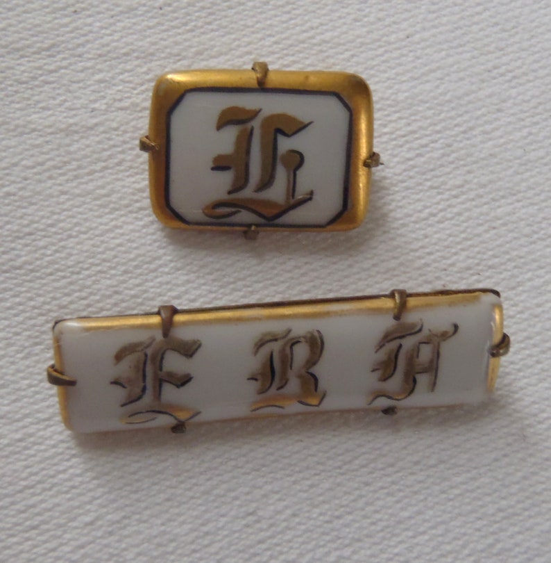May include: Two gold-rimmed porcelain brooches with black and gold lettering. The first brooch is square and features the letter "F" in a stylized font. The second brooch is rectangular and features the letters "F R M" in a stylized font.