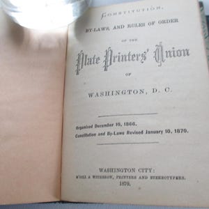 May include: An open book displaying the title page of the "Constitution, By-Laws, and Rules of Order of the Plate Printers' Union of Washington, D.C." Published in 1870. The text is in black ink on aged paper.