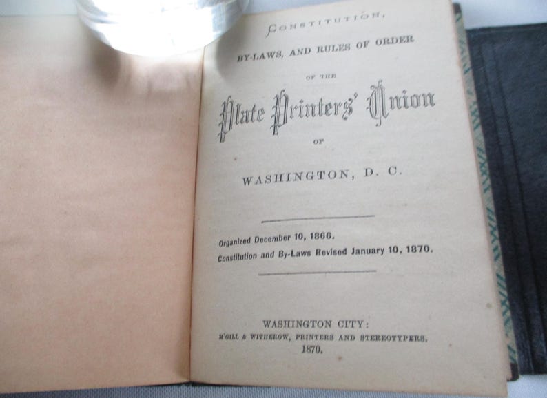 May include: An open book displaying the title page of the "Constitution, By-Laws, and Rules of Order of the Plate Printers' Union of Washington, D.C." Published in 1870. The text is in black ink on aged paper.